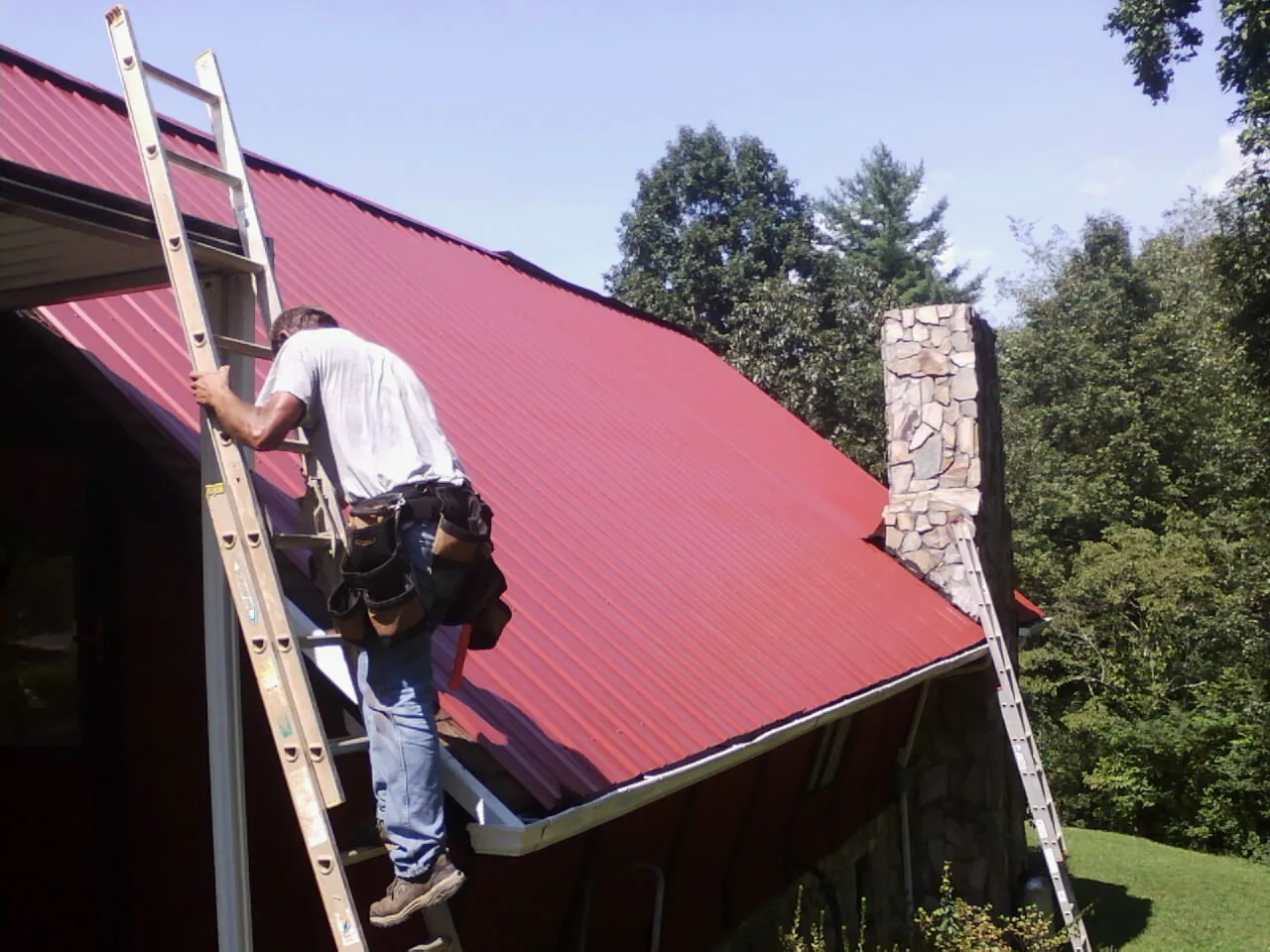 Roofer on a ladder beside a red metal roof and stone chimney — Soffit & Fascia Repair by Roofing Brothers in Camden Wyoming